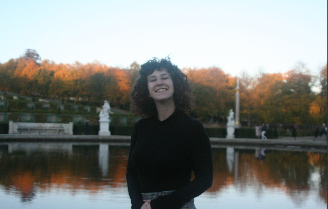 Adri, a female presenting person in a black turtleneck with curly brown hair, is standing in front of a lake and colorful autumn forest. She is smiling towards the camera.